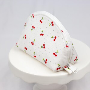 May include: White zippered pouch with a cherry print and white polka dots. The pouch is on a white cake stand. The text "sugar apple seams" is on a green circle.