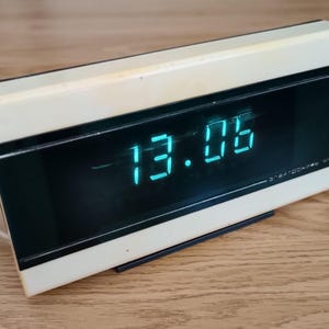 Puede incluir: Un reloj digital vintage con una parte superior de color crema y un área de visualización negra. La hora que se muestra es 13:06 en dígitos de color verde azulado brillante. El reloj tiene forma rectangular y está sobre una superficie de madera.