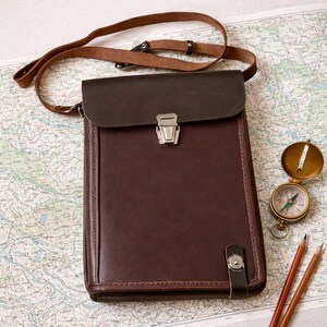 May include: A dark brown leather messenger bag with a rectangular flap, a silver metal clasp, and an adjustable shoulder strap. The bag features a vertical leather tab with a silver snap button closure at the bottom right corner of the front panel.