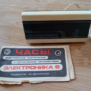May include: A retro electronic table clock with a cream-coloured top and a black front panel. The clock has a white power cord and a paper manual with Cyrillic text. The manual reads "ELECTRONIKA 6".