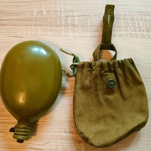 May include: An olive green military-style canteen with a matching canvas pouch. The canteen has a rounded, oval shape and a screw-on cap. The pouch has a drawstring closure and a button detail. The canteen and pouch are made of durable materials.