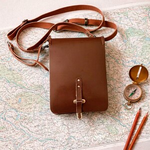 May include: A brown leather vertical crossbody bag with a long adjustable strap and a central buckle closure. Beside the bag sits a round brass compass with a green needle and two sharpened wooden pencils, all resting on a detailed topographic map.