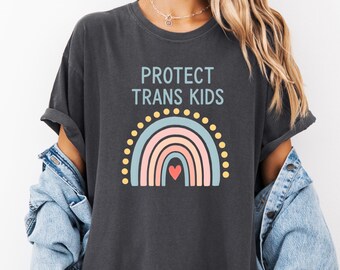 Puede incluir: Camiseta gris oscuro con el texto "PROTECT TRANS KIDS" sobre un diseño de arcoíris. El arcoíris presenta arcos celeste, rosa y melocotón con un corazón rojo en el centro. Puntos amarillos bordean la parte superior del arcoíris.