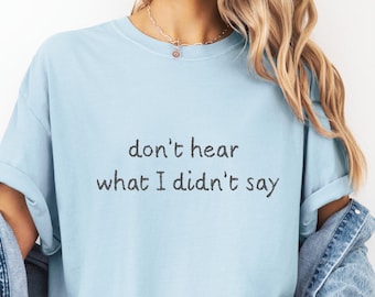 May include: Light blue t-shirt with the text "don't hear what I didn't say" in black. The shirt has short sleeves and a relaxed fit. The model is wearing a denim jacket and a silver necklace.