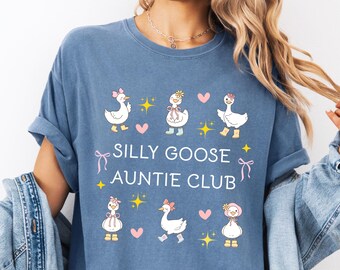 Może przedstawiać: Niebieska koszulka z napisem "SILLY GOOSE AUNTIE CLUB" i ilustracjami kreskówkowych gęsi z kokardkami, kwiatami i sercami. Koszulka ma krótkie rękawy i luźny krój.