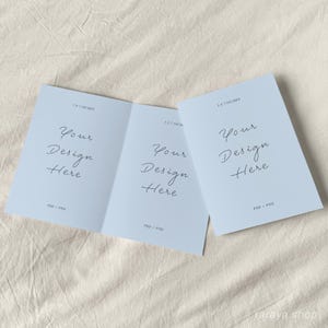 May include: A folded card mockup with a light blue background. The card is 5.5 x 4.25 inches and has the text "Your Design Here" on each side. The card is lying on a light beige fabric surface.