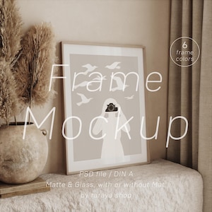 May include: A mockup of a frame with a light brown background and a white silhouette of a person holding a camera. The frame is on a light brown surface with a vase of dried pampas grass in the foreground. The text "Frame Mockup" is superimposed on the image. The text "PSD file / DIN A" is below the frame. The text "Matte & Glass, with or without Mat." is below the previous text. The text "by raraya shop" is below the previous text.