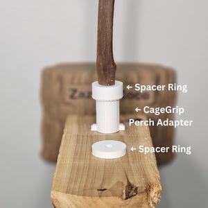 Puede incluir: Primer plano de una percha de madera con una rama marrón y accesorios blancos. Los accesorios incluyen un adaptador de percha CageGrip y dos anillos espaciadores. La percha está montada sobre una base de madera.