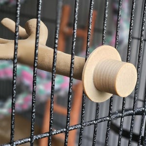 Puede incluir: Un posadero para pájaros de color marrón claro con diseño de rama, fijado a una jaula negra. El posadero tiene un extremo circular y una superficie texturizada, proporcionando una sensación natural a las aves. Las barras de la jaula son visibles.