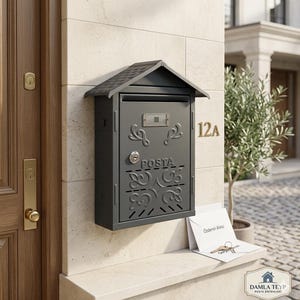 Könnte beinhalten: Ein dunkelgrauer Briefkasten mit kleinem Dach, montiert an einer hellen Steinmauer. Der Briefkasten hat dekorative Schnörkel und das Wort "POSTA" ist sichtbar. Die Adresse "12A" ist in goldenen Buchstaben.