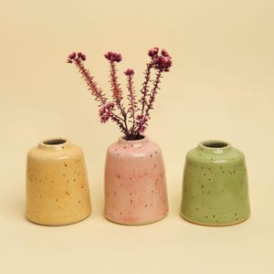 May include: Three small ceramic vases in yellow, pink, and green, each with a speckled finish. The pink vase holds a sprig of purple flowers. The vases have a rounded shape and a narrow opening, ideal for small floral arrangements.
