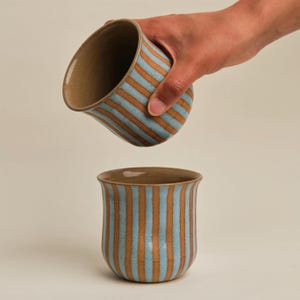 May include: Two ceramic mugs with a light blue and brown striped pattern. One mug is held by a hand, tilted as if pouring. The other mug is upright. Both mugs have a brown interior and a rounded shape.