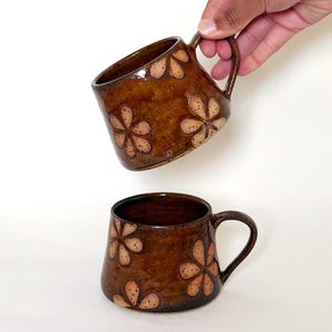 May include: Two ceramic mugs with a glossy, speckled brown glaze and a repeating light-coloured flower pattern. Each mug features a wide, rounded body, a sturdy handle, and a smooth interior finish, showcasing a consistent handcrafted aesthetic.