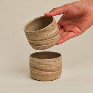 May include: Two handmade ceramic cups with a natural, earthy aesthetic. The cups feature a unique, swirling pattern in shades of beige and brown. One cup is held in the air, while the other sits below. The cups are ideal for coffee, tea, or other beverages.