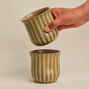 May include: Two ceramic mugs with vertical stripes in light green and brown. One mug is held by a hand, tilted slightly above the other mug. The mugs have a rounded base and a slightly flared rim.