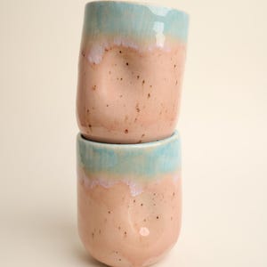 May include: Two stacked ceramic mugs with a light blue and pink speckled glaze. The mugs have a unique, slightly uneven shape.