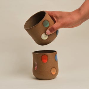 May include: Two brown ceramic mugs with colourful polka dots. One mug is held by a hand, tilted slightly, while the other sits upright below. The dots are in various colours, including red, blue, green, and orange.