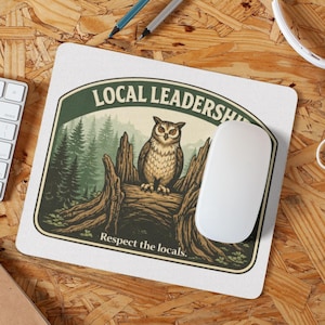 May include: A square mouse mat featuring an owl illustration. The design shows an owl on a tree stump in front of a forest scene, with the text "LOCAL LEADERSHIP" and "Respect the locals." A white mouse and keyboard are visible.