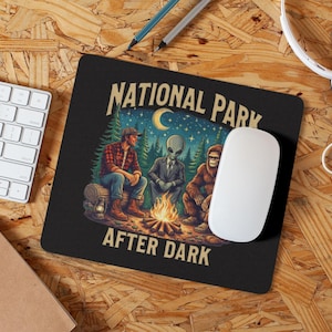 May include: Black mousepad with the text "NATIONAL PARK AFTER DARK" featuring a campfire scene with a man, an alien, and Bigfoot. The mousepad is rectangular and has a white mouse on top.