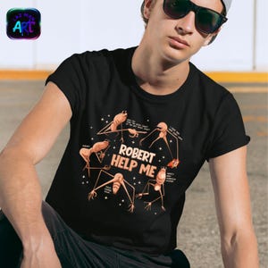 Könnte beinhalten: Schwarzes T-Shirt mit einer humorvollen Grafik, die insektenartige Figuren und den Text "ROBERT HELP ME" zeigt. Das Design umfasst Sprechblasen und Sternakzente, die eine verspielte und skurrile Ästhetik erzeugen. Das Shirt hat einen klassischen Rundhalsausschnitt.