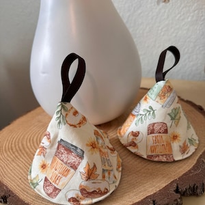 May include: Two cream-colored, cone-shaped fabric ornaments with brown ribbon loops. The ornaments feature a fall-themed print with coffee cups, donuts, leaves, and the text "AUTUMN VIBES". They rest on a wooden slice.