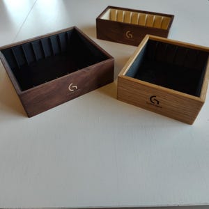 May include: Three wooden jewelry boxes in varying shades of brown. Each box has a dark interior and a logo with the letter "G" and the words "Custom Gems". The boxes are rectangular and appear to be made of wood.