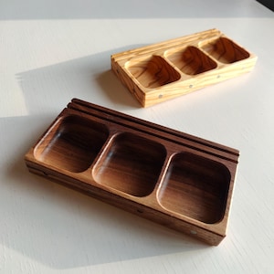 Könnte beinhalten: Zwei Holzserviertabletts mit jeweils drei Fächern. Eines ist aus hellem Holz mit sichtbarer Maserung, das andere aus dunklerem Holz. Beide rechteckigen Tabletts sind zum Servieren von Snacks oder kleinen Gegenständen konzipiert.