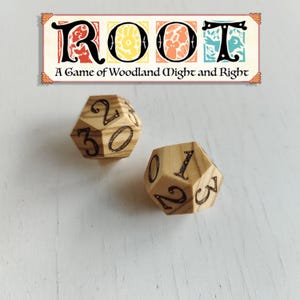 May include: Two wooden, multi-sided dice with black numbers are displayed. The dice are numbered 0-3. Above the dice is a sign that reads "ROOT A Game of Woodland Might and Right" in a decorative font with colorful accents.