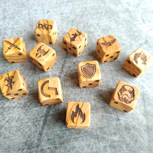 Peut inclure: Douze dés en bois avec différents symboles et motifs, dont une épée, un bouclier, un dragon, un feu, un personnage de Star Wars et les lettres "D&D".