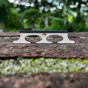 May include: A wooden banjo bridge featuring a light-colored base with two oval cutouts and a dark-colored top edge. The letters CRB are clearly printed in black on the front center of the light wood section of the bridge component.