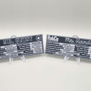 May include: Two black metal nameplates with white text. The top plate reads "RobCo Industries" and "Mr. Gutsy" with the text "Made in the U.S.A." below. The bottom plate reads "RobCo Industries" and "Mr. Handy" with the text "How may I serve you?" below. Both plates have a star in the top right corner and list model number, manufacture date, production facility, preferred name, personality class, power unit, serial number, QC test date, design spec, max carry weight, unit weight, and power output.