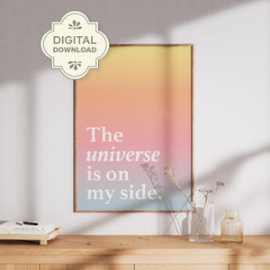 May include: A framed print with a gradient background of yellow, pink, and blue. The white text reads "The universe is on my side." A "DIGITAL DOWNLOAD" sticker is in the upper left corner. The print is displayed on a wooden surface.