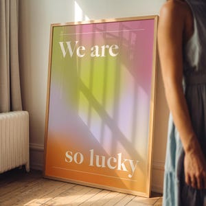 Może przedstawiać: Oprawiony wydruk artystyczny z gradientowym tłem w odcieniach pomarańczu, żółci, różu i fioletu. Słowa "We are so lucky" są wydrukowane na biało. Rama jest w jasnobrązowym kolorze.