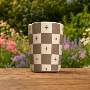 Puede incluir: Un vaso cil&iacute;ndrico de cer&aacute;mica con un patr&oacute;n de tablero de ajedrez repetitivo en tonos alternos de beige mate y marr&oacute;n apagado, con cada cuadrado acentuado por un peque&ntilde;o icono de estrella dorada de cuatro puntas centrado.