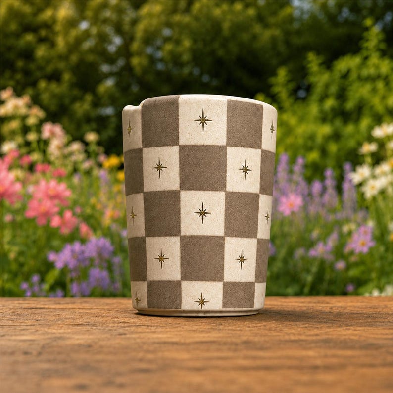 Puede incluir: Un vaso cil&iacute;ndrico de cer&aacute;mica con un patr&oacute;n de tablero de ajedrez repetitivo en tonos alternos de beige mate y marr&oacute;n apagado, con cada cuadrado acentuado por un peque&ntilde;o icono de estrella dorada de cuatro puntas centrado.