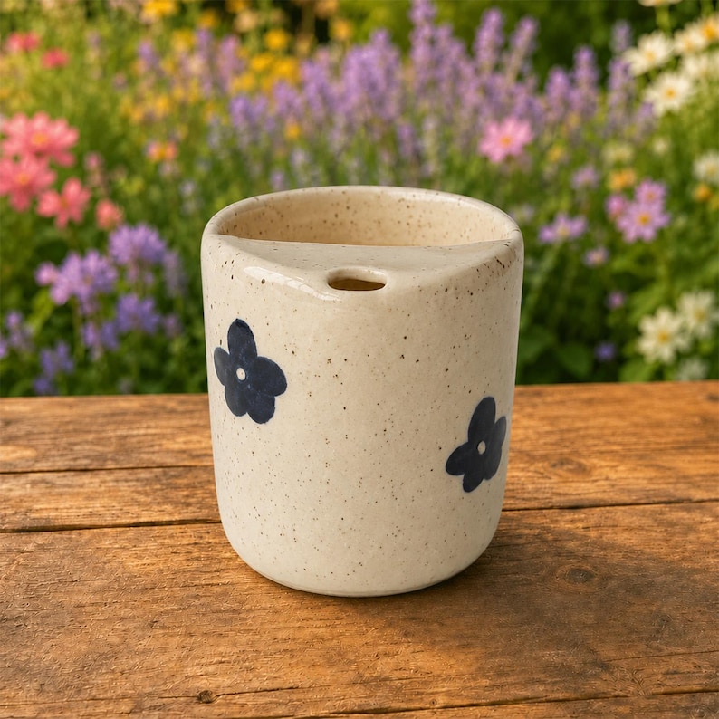 May include: A cylindrical ceramic cup with a speckled cream finish and a small circular opening near the top rim. The exterior features hand-painted dark blue five-petal flower motifs distributed across the surface of the vessel.