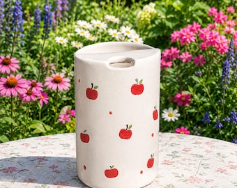 Op de afbeelding: Een cilindrische keramische keukengereihouder in een gebroken witte kleur, versierd met een herhalend patroon van kleine rode appels en kleine rode stippen. De bovenkant heeft een halfronde opening en een glad, glanzend oppervlak.