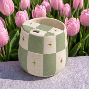 Puede incluir: Un vaso de cerámica con un patrón de cuadros en color crema y verde salvia moteado. La superficie presenta pequeños detalles de estrellas doradas y una tapa con una pequeña abertura. El recipiente tiene una forma cilíndrica y mate.