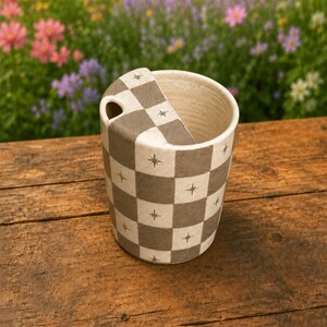 Puede incluir: Un vaso cil&iacute;ndrico de cer&aacute;mica con un patr&oacute;n de tablero de ajedrez en tonos alternos de beige y gris apagado. Cada cuadrado gris est&aacute; decorado con un peque&ntilde;o icono de estrella dorada. El vaso incluye una tapa parcial con una peque&ntilde;a abertura.