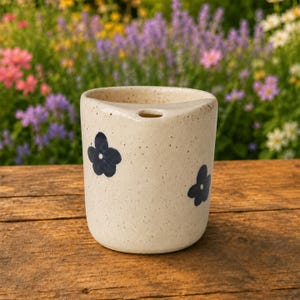 May include: A cylindrical ceramic cup with a speckled cream-colored finish, featuring two dark blue hand-painted flower motifs on the exterior and a small circular hole near the top rim, designed for holding items or drinking.
