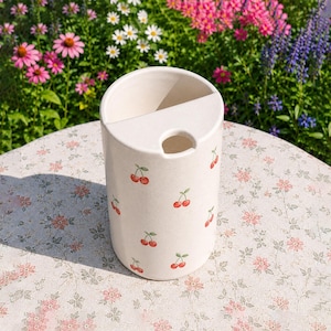 May include: A cylindrical off-white ceramic utensil holder featuring a repeating pattern of small red cherries with green stems. The top surface has a semi-circular opening and a smaller circular hole, providing a functional design for kitchen tools.