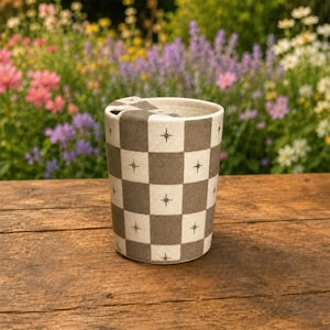 Puede incluir: Un vaso cil&iacute;ndrico de cer&aacute;mica con un patr&oacute;n de tablero de ajedrez de cuadrados alternos en color blanco roto y gris apagado. Cada cuadrado gris est&aacute; decorado con un peque&ntilde;o icono de estrella oscura de cuatro puntas. El recipiente tiene una tapa plana.