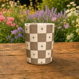 Puede incluir: Una taza cil&iacute;ndrica de cer&aacute;mica con un patr&oacute;n de tablero de ajedrez repetitivo en tonos alternos de beige y gris oscuro. Cada cuadrado est&aacute; decorado con un peque&ntilde;o icono de estrella de cuatro puntas centrado. La taza tiene un acabado liso.