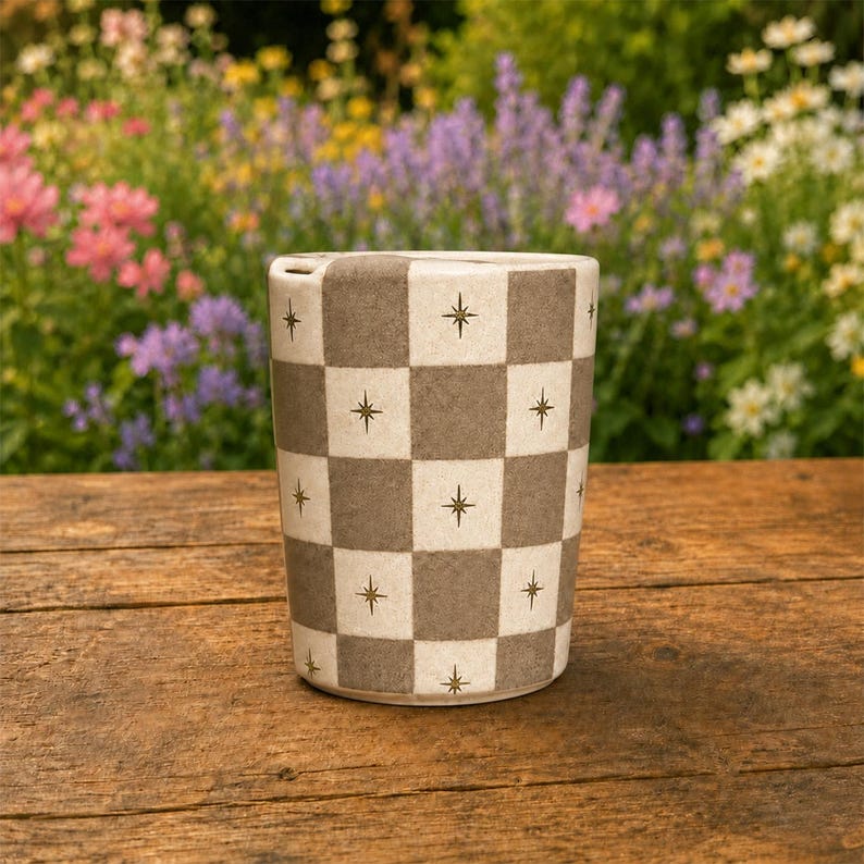Puede incluir: Una taza cil&iacute;ndrica de cer&aacute;mica con un patr&oacute;n de tablero de ajedrez repetitivo en tonos alternos de beige y gris oscuro. Cada cuadrado est&aacute; decorado con un peque&ntilde;o icono de estrella de cuatro puntas centrado. La taza tiene un acabado liso.