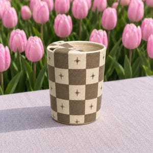 Peut inclure: Un gobelet cylindrique en céramique avec un motif à damier marron et crème. Chaque carré marron est orné d'une petite étoile dorée à quatre branches. Le gobelet possède un couvercle en céramique assorti avec une petite ouverture circulaire.