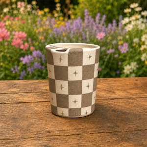 Puede incluir: Un vaso cil&iacute;ndrico de cer&aacute;mica con un patr&oacute;n de tablero de ajedrez en tonos alternos de beige y marr&oacute;n apagado. Cada cuadrado est&aacute; decorado con un peque&ntilde;o icono de estrella de color dorado. El vaso tiene un borde liso y una base plana.