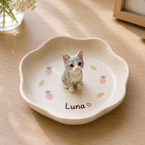 Puede incluir: Un plato de cerámica beige con forma de flor que presenta una pequeña figura tridimensional de un gatito atigrado gris en el centro. El plato está decorado con pequeñas ilustraciones de fresas rosas y hojas verdes, con el nombre Luna y un corazón.