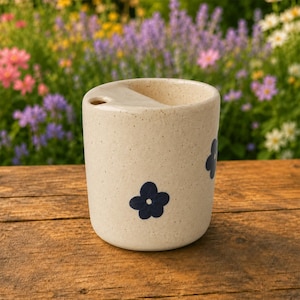 May include: A cylindrical ceramic cup with a speckled cream-colored finish and a small circular opening on the top rim. The exterior is decorated with several dark blue, five-petaled flower motifs arranged around the body of the vessel.