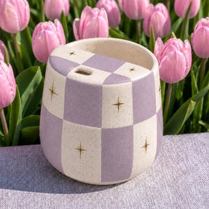 May include: A ceramic tumbler with a speckled cream and lavender checkered pattern, featuring small gold starburst icons on the cream squares and a small drinking hole on the top rim, set against a background of vibrant pink tulip flowers.