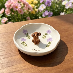 May include: A small, off-white ceramic dish with a brown dog figurine in the center. The dish is decorated with purple flowers and green leaves. The word "Momo" is written on the dish.